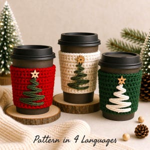 Könnte beinhalten: Drei schwarze Einweg-Kaffeebecher mit gehäkelten Manschetten in Rot, Creme und Dunkelgrün. Jede Manschette zeigt ein aus Kordel gefertigtes Weihnachtsbaum-Motiv mit einem kleinen Holzstern. Der Text "Pattern in 4 Languages" ist zu sehen.