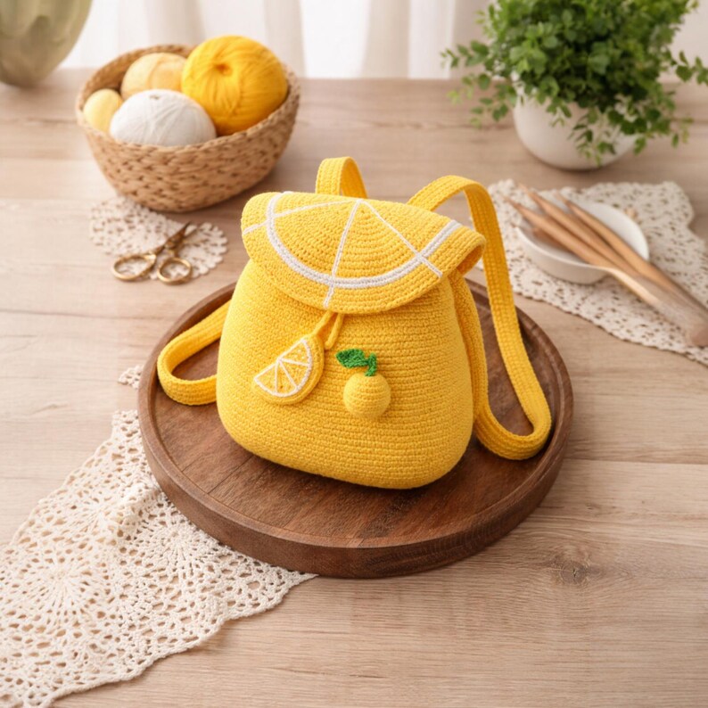 May include: A bright yellow crochet backpack featuring a flap designed to look like a citrus slice with white segment lines. The bag includes two shoulder straps, a small dangling crochet citrus slice charm, and a tiny green leaf accent.