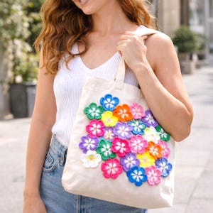 May include: A cream-colored tote bag adorned with a cluster of colorful, crocheted flowers. The flowers are in shades of blue, pink, purple, yellow, orange, and white. The bag has a simple design with a rectangular shape and sturdy handles.
