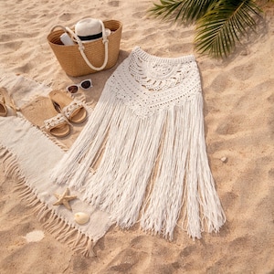 May include: A white crochet skirt with long fringe, on a sandy beach. A straw tote bag, sunglasses, sandals, and a beach towel are also visible. The skirt has a detailed, openwork design at the waist.