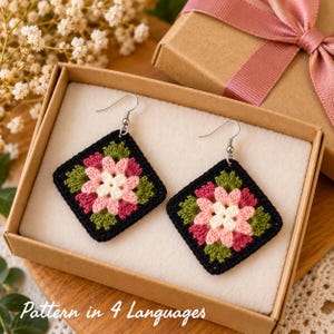 Puede incluir: Un par de pendientes cuadrados de ganchillo con un motivo floral en tonos rosa, blanco y verde, bordeados con hilo negro. Los pendientes están sujetos a ganchos de metal plateado y se presentan dentro de una caja de regalo de cartón.