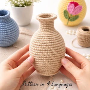 Può includere: Un vaso beige all'uncinetto è tenuto al centro dell'immagine. Il vaso ha un corpo arrotondato e un collo stretto. Altri vasi all'uncinetto blu e gialli sono sullo sfondo. Il testo "Pattern in 4 Languages" è in basso.