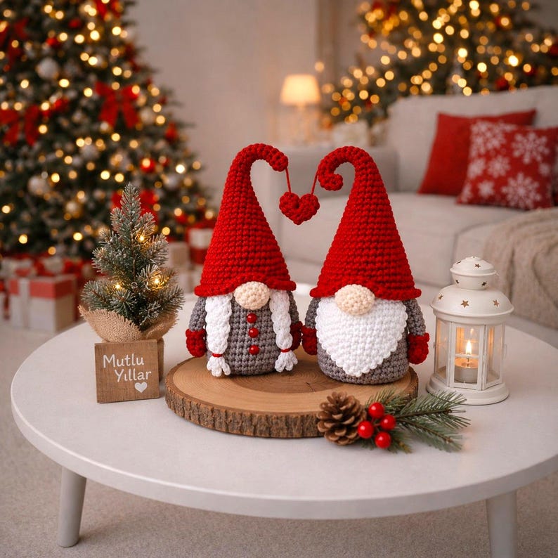 Peut inclure: Deux gnomes en crochet avec des chapeaux rouges et des tenues grises, reli&eacute;s par un ornement en forme de c&oelig;ur. Ils sont pos&eacute;s sur une tranche de bois, avec un petit sapin de No&euml;l et une lanterne. Le texte sur le panneau en bois dit "Mutlu Yillar".
