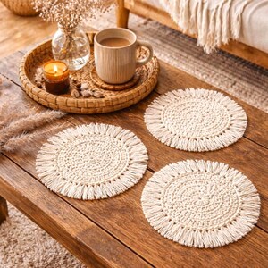 May include: Three cream-colored, round macrame coasters with fringed edges sit on a rustic wooden table. A woven tray holds a mug of coffee, a candle, and a small vase with dried flowers. The scene evokes a cozy, bohemian aesthetic.
