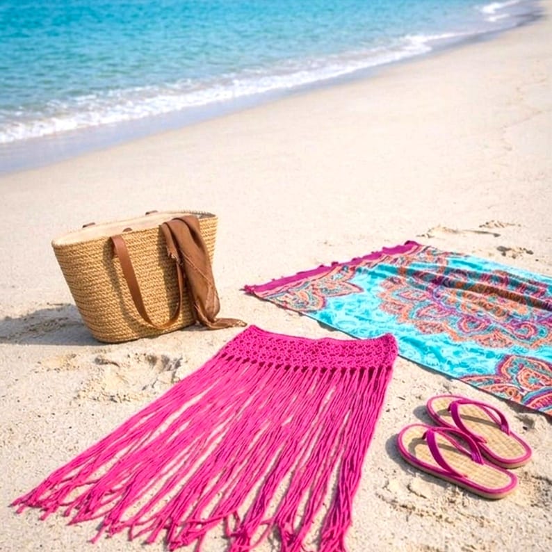 Pode incluir: Uma cena de praia com uma bolsa tote tecida, uma toalha de praia estampada, uma saia com franjas rosa brilhante e um par de chinelos rosa. Os itens est&atilde;o dispostos em uma praia arenosa com o oceano ao fundo.