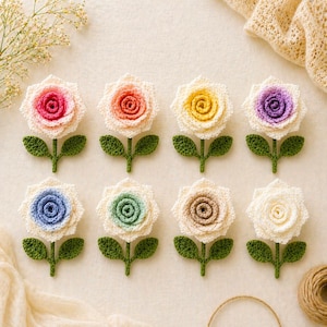 May include: Eight handmade crochet flowers in various colors, including pink, orange, yellow, purple, blue, green, brown, and white. Each flower has a green stem and leaves. The flowers are arranged on a cream-colored surface.