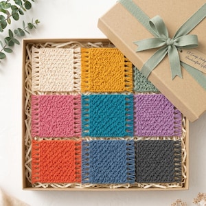 Op de afbeelding: Een geschenkdoos met negen vierkante gehaakte onderzetters in verschillende kleuren. De kleuren zijn cr&egrave;me, geel, roze, turquoise, paars, oranje, blauw en grijs, elk met gefranjerde randen. De doos is vastgebonden met een groen lint.