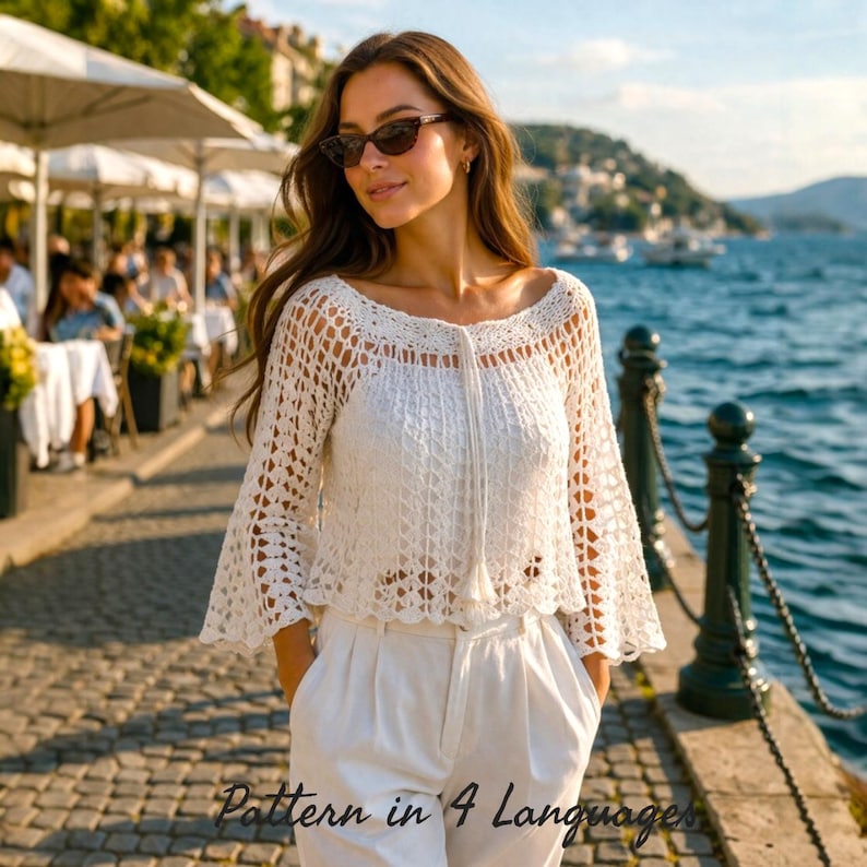 Cream crochet top worn by a woman standing outdoors in a sunlit park, featuring an airy lace mesh pattern, wide boat neckline, and loose bell sleeves. The handmade crochet blouse has a relaxed fit with delicate stitch details and a soft drape. Styled with beige high-waisted pants, this lightweight summer top pattern is ideal for beginners and includes step-by-step instructions. Perfect for boho outfits, beachwear, and everyday casual fashion.