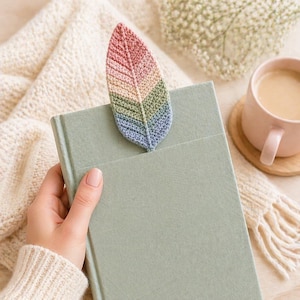 May include: A handmade, crochet leaf bookmark in shades of pink, yellow, green, and blue, inserted into a light green book. The leaf-shaped bookmark is made of yarn and has a detailed, textured appearance. A pink mug of coffee sits on a wooden coaster nearby.