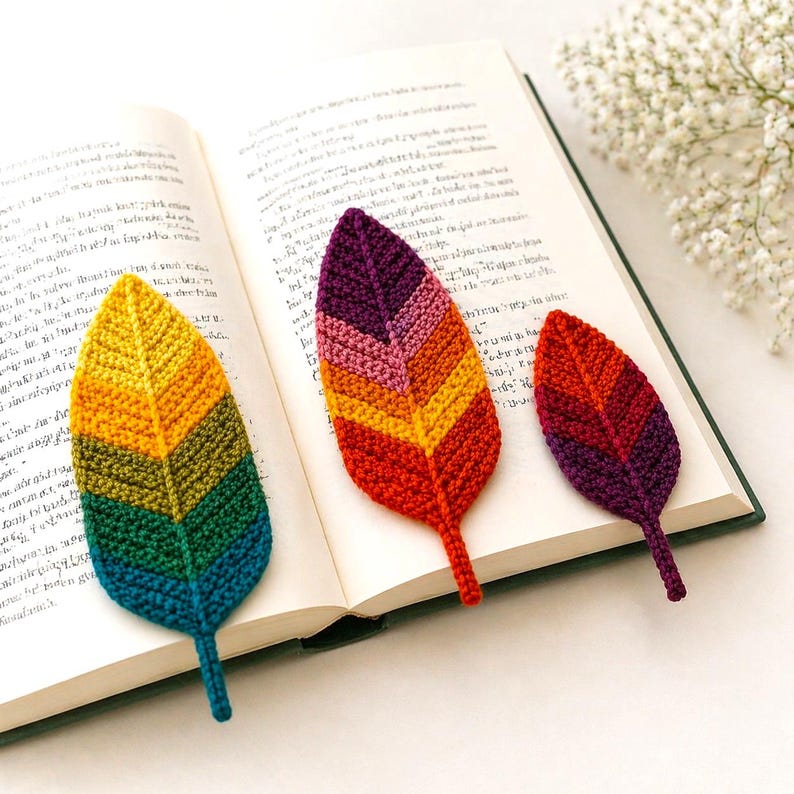 May include: Three handmade crochet leaf bookmarks in vibrant colors. One is yellow to blue, another is red to purple, and the third is a smaller version of red to purple. The bookmarks are placed on the open pages of a book.