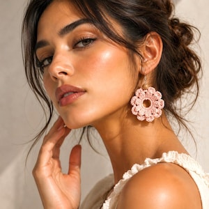 May include: A close-up of a person wearing a pair of pink crochet flower earrings. The earrings are a light pink color and have a circular design with a wooden bead at the top. The person is wearing a cream-colored top.