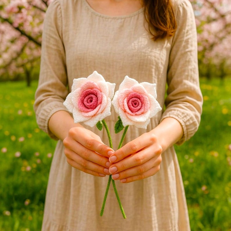 May include: Two handmade crochet roses with pink and white petals and green stems are held in front of a person wearing a beige dress. The roses have a detailed, textured appearance. The background is a blurred view of a garden with pink blossoms and green grass.