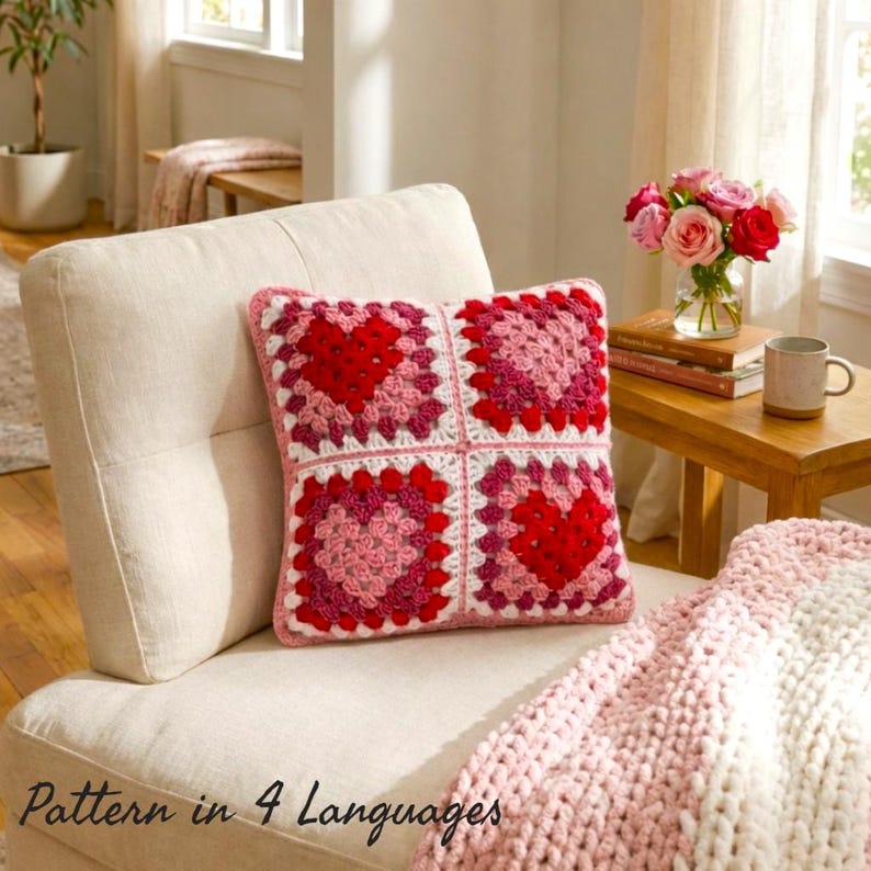 Cozy living room scene featuring a handmade crochet pillow with heart granny square motifs in red, pink and white, placed on a cream armchair. Styled with a soft knitted blanket, wooden side table and fresh flowers, creating a warm romantic home decor aesthetic. Perfect for Valentine’s Day decor and crochet lovers. Pattern available in 4 languages: English, German, French and Spanish