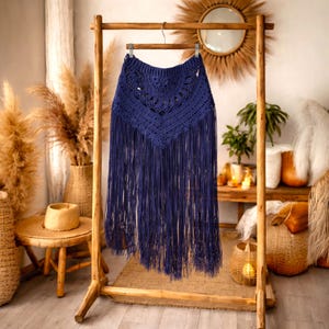 May include: A navy blue crochet skirt with long fringe detail. The skirt is displayed on a wooden clothes rack. The top section has a detailed crochet pattern, and the fringe hangs down, creating a flowing effect. The skirt is a bohemian style.