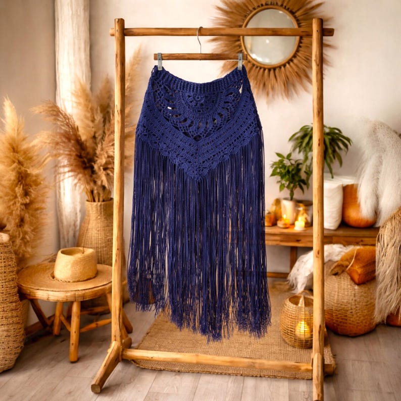 May include: A navy blue crochet skirt with long fringe detail. The skirt is displayed on a wooden clothes rack. The top section has a detailed crochet pattern, and the fringe hangs down, creating a flowing effect. The skirt is a bohemian style.