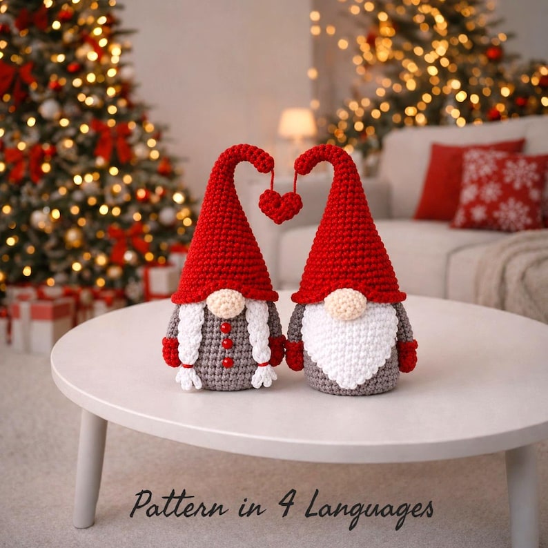 Festive crochet gnome couple on a white table in a cozy Christmas living room. Handmade amigurumi gnomes wear tall red hats with a hanging heart detail, one with braided hair and one with a fluffy white beard. Warm Christmas tree lights, wrapped gifts, and soft holiday décor create a cozy bokeh background. Text reads “Pattern in 4 Languages,” promoting a multilingual crochet PDF pattern for Christmas décor or handmade gift ideas.