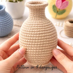 Puede incluir: Un jarrón beige tejido a ganchillo con un cuerpo redondeado y un cuello acampanado. El jarrón está sostenido por dos manos. El texto "Pattern in 4 Languages" está en la parte inferior de la imagen. Otros artículos tejidos a ganchillo están en el fondo.