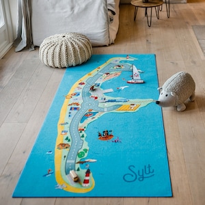 May include: A blue and yellow play mat with a map of the island of Sylt, Germany. The mat features a variety of illustrations, including houses, cars, boats, and a lighthouse. A small stuffed hedgehog is sitting on the mat.