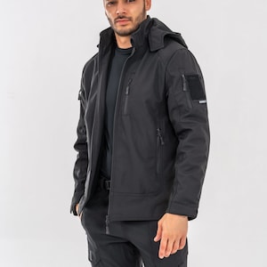 May include: Black tactical jacket with a hood, multiple zippered pockets, and a patch on the upper arm. The jacket is paired with black cargo pants. The model is standing in front of a white background.