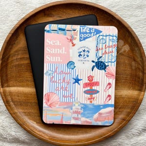 Puede incluir: Un dispositivo lector electrónico rectangular con una pegatina decorativa que presenta un collage de ilustraciones con temática de playa, incluyendo un faro, conchas, una tortuga marina y el texto "Life is good!", "Sea. Sand. Sun." y "Saltwater heals everything."