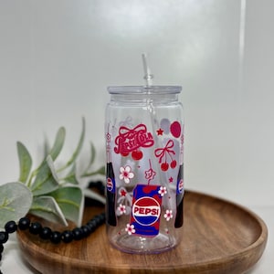 May include: Clear glass tumbler with a lid and straw, decorated with a Pepsi design. The design includes the Pepsi logo, cherries, flowers, and the Pepsi Cola logo in red and white. The tumbler is sitting on a wooden tray.