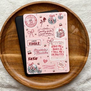 May include: A black e-reader device covered with a light pink adhesive skin featuring various illustrations including disco balls, cherries, bows, a rose, and stacks of books. Text includes "Kindle Girlie," "pretty girls read books," and "Buy Me Books & Tell Me I'm Pretty."