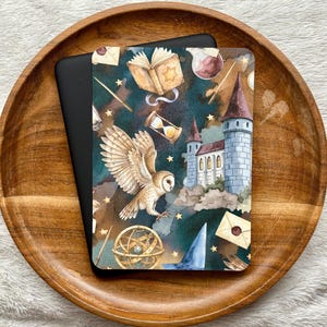 May include: A rectangular e-reader cover featuring a dark teal background with a pattern of a flying barn owl, a stone castle with towers, an open book, an hourglass, a golden armillary sphere, a blue pointed hat, and sealed envelopes.
