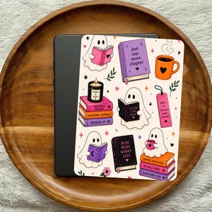 May include: A black e-reader device featuring a cream-colored skin with a repeating pattern of white ghosts reading books, stacks of colorful books with titles like 'Romance Reader' and 'Read More Worry Less', an orange mug, and a pink bookmark.
