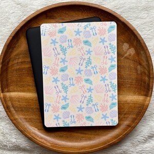 Puede incluir: Una pegatina rectangular con un patrón repetitivo de conchas, estrellas de mar, corales y pequeños lazos en tonos pastel sobre un fondo blanco. La pegatina está colocada sobre un dispositivo rectangular negro, ambos sobre una bandeja de madera.