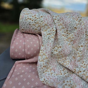 May include: Two rolls of fabric are displayed. One roll features a light pink background with a repeating pattern of small white four-petaled flowers. The other roll showcases a cream background with a dense floral print of yellow flowers.
