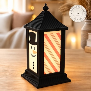 May include: A black lantern with a peaked roof and a square base. One side features a snowman graphic with a black top hat and orange nose, while the adjacent side displays red and white diagonal stripes. A circular badge reads Digital Download STL + 3MF.