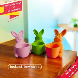 Op de afbeelding: Drie 3D-geprinte eierrekjes in de vorm van konijnen in roze, groen en oranje. De konijnen-eierrekjes staan op een houten tafel. De tekst "Direct downloaden" is zichtbaar in de afbeelding. De tekst "Printinformatie inbegrepen!" is zichtbaar in de afbeelding.