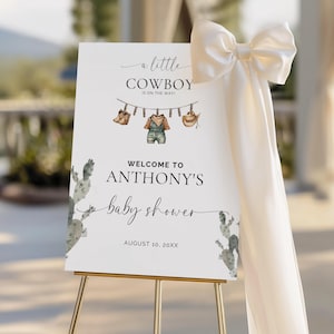 May include: A baby shower welcome sign on a gold easel. The sign reads "a little COWBOY is on the way!" with illustrations of cowboy boots, overalls, and a hat. The sign welcomes guests to Anthony's baby shower.