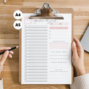 Può includere: Un foglio di pianificazione giornaliera tenuto su una lavagna di legno. Il pianificatore ha un design rosa e nero con sezioni per fasce orarie, priorità, affermazioni e note. Il testo "Aperçu du jour" è in alto.