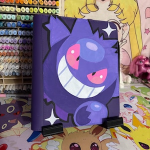 Puede incluir: Una carpeta morada con una gran ilustración pintada a mano del personaje Gengar. El diseño muestra a la criatura redonda de color morado oscuro con una amplia sonrisa, ojos rosados y dos estrellas blancas de cuatro puntas sobre una superficie.