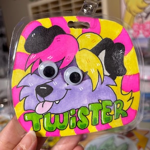 May include: A handmade badge featuring a cartoon dog with googly eyes, purple fur, and a pink tongue. The badge is outlined in black and has the name "TWISTER" in green letters. The background is yellow and pink striped.