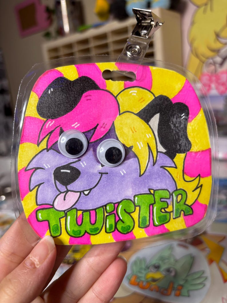May include: A handmade badge featuring a cartoon dog with googly eyes, purple fur, and a pink tongue. The badge is outlined in black and has the name "TWISTER" in green letters. The background is yellow and pink striped.