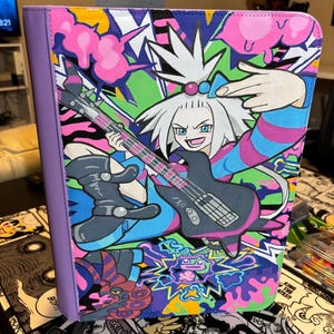 May include: A purple-spined binder featuring a vibrant, multi-colored graphic illustration of a spiky-haired character playing a black electric guitar. The artwork includes bold pink, green, and blue graffiti-style accents and abstract shapes.