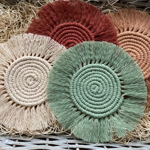 May include: Four handmade crocheted coasters with fringe. The coasters are in shades of beige, green, brown, and rust.