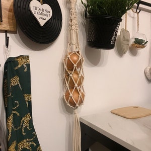 May include: A white macrame onion holder with three onions hanging inside. The holder is hanging from a black metal rod on a white wall. There is a black and white sign that says "I'll Do It Now in a Minute!" hanging on the wall to the left of the holder.