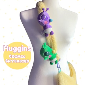 May include: A yellow fuzzy shoulder bag with a purple and green alien plush toy attached to the strap. The bag has the text "Huggins Cosmic Crybabies" on it.