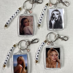 May include: Four keyrings with clear acrylic photo inserts featuring a woman's image. Each keyring has a beaded strand spelling "SABRINA", a star charm, and a silver-tone clasp. The photos are in colour and black and white.