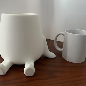 May include: A white ceramic planter with four legs shaped like feet. The planter is next to a plain white ceramic mug.