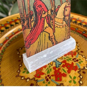 May include: A white selenite crystal stand with a tarot card featuring a red-robed figure riding a white horse. The card is titled "The Chariot" and features a colorful background.