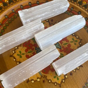 May include: Five selenite sticks, also known as selenite wands, are arranged on a wooden plate with a colorful floral design. The sticks are white and have a striated texture.