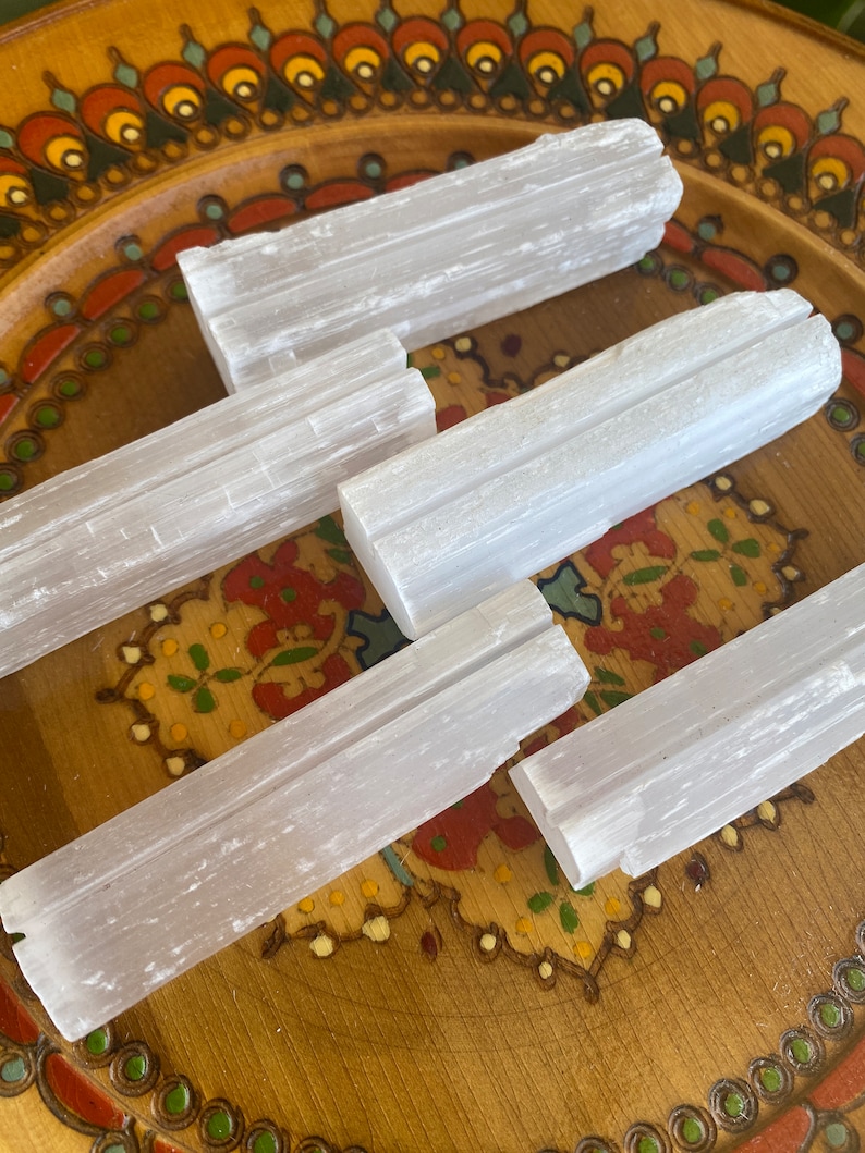 May include: Five selenite sticks, also known as selenite wands, are arranged on a wooden plate with a colorful floral design. The sticks are white and have a striated texture.