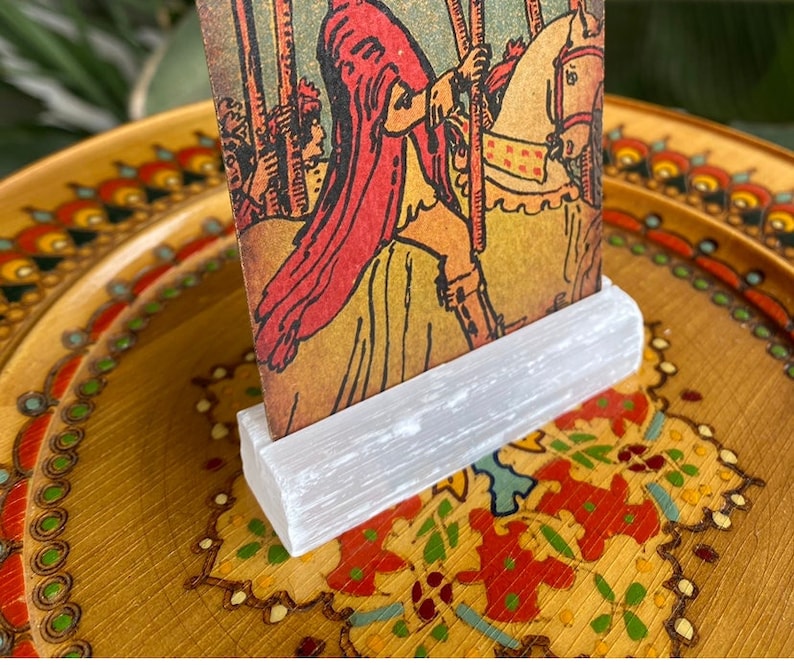 May include: A white selenite crystal stand with a tarot card featuring a red-robed figure riding a white horse. The card is titled "The Chariot" and features a colorful background.