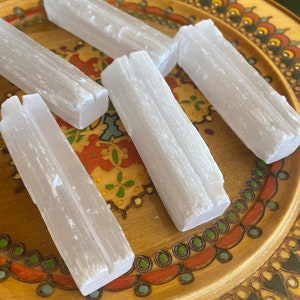 May include: Five white selenite sticks on a wooden plate with a colorful floral design. The sticks are smooth and have a natural, crystalline texture.
