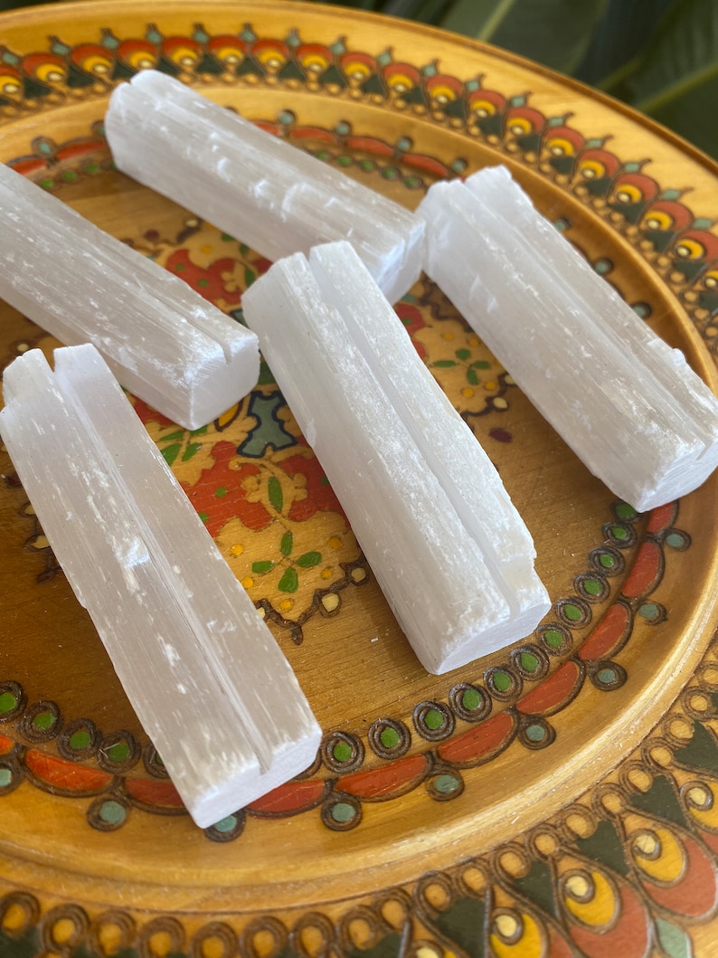 May include: Five white selenite sticks on a wooden plate with a colorful floral design. The sticks are smooth and have a natural, crystalline texture.