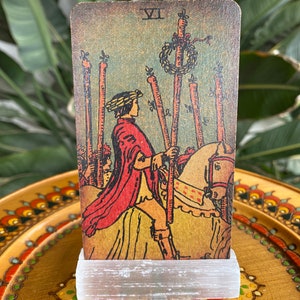 May include: A vintage tarot card with a worn, aged look. The card depicts a man on horseback, holding a staff. The card number VII is visible in the top right corner. The card is on a white stand.